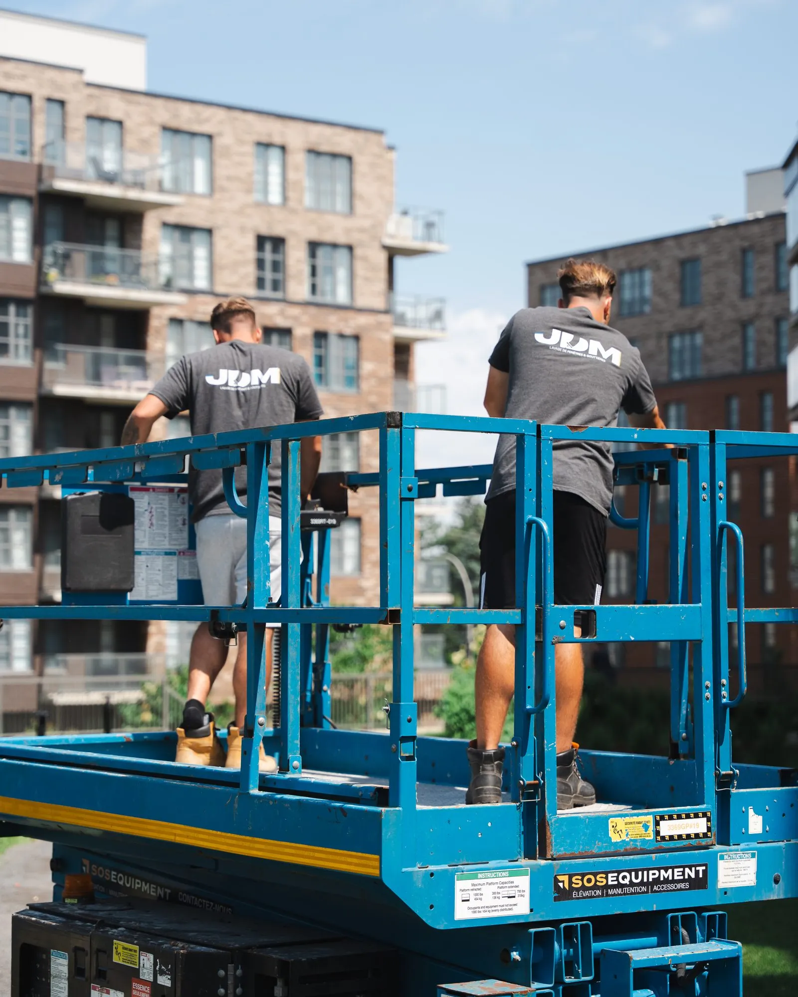 Deux techniciens de Lavage JDM travaillent depuis une nacelle élévatrice devant la façade vitrée d'un immeuble à condos.